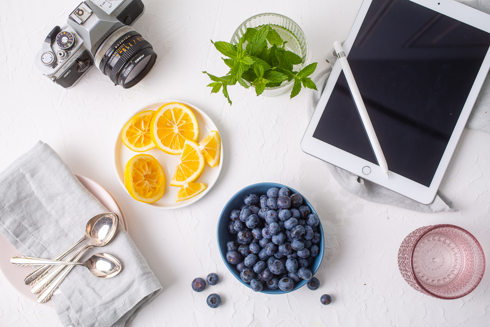 Bowl of Blueberries, lemons on a plate, glass of water next to an iPad and a camera